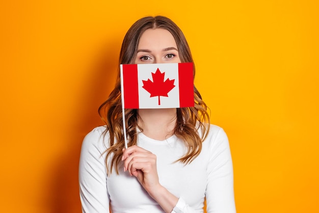 Studentin bedeckt ihr Gesicht mit einer kleinen Kanada-Flagge isoliert über orangefarbenem Hintergrund, Kanada-Tag, Feiertag, Jahrestag der Konföderation, Kopierraum