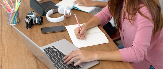 Studentin der Universität, die Notiz auf leerem Notizbuch beim Suchen in Formation auf Laptop in Bibliothek nimmt