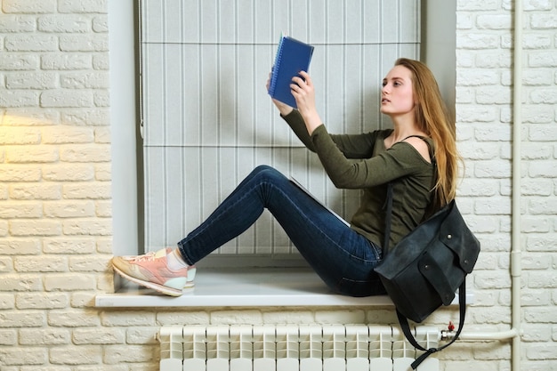 Studentin, die auf der Fensterbank sitzt, studiert, Notizbuch liest. Junge schöne Frau mit Rucksack-Studentin, Bildung, Selbststudium