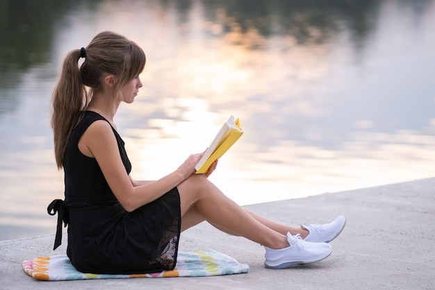 Studentin, die draußen im Sommerpark sitzt und Lehrbuch liest. Bildungs- und Studienkonzept.