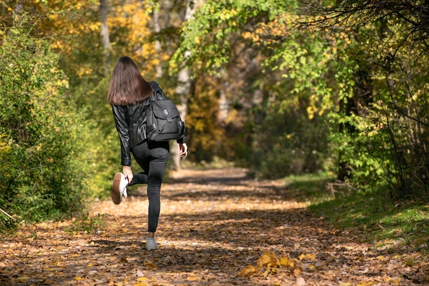 Studentin geht im Herbstpark. Mädchen mit dunklen Haaren glättet ihre Schuhe. Einsam durch den Wald gehen. Rückansicht.