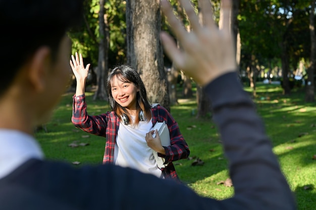 Studentin grüßt oder verabschiedet sich von ihrer Freundin im Park und winkt mit der Hand auf Wiedersehen Geste