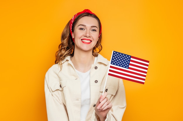 Studentin hält eine kleine amerikanische Flagge und lächelt isoliert über orange Hintergrund