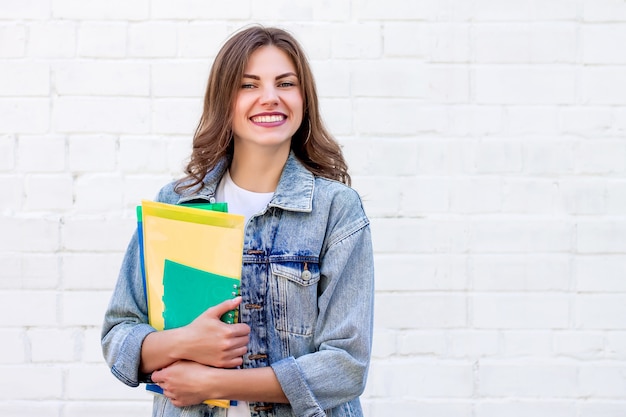 Studentin hält Ordner und ein Notizbuch in ihren Händen und lächelt auf einem Hintergrund einer weißen Backsteinmauer, Kopienraum