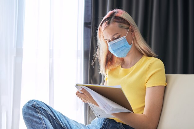 Studentin in medizinischen Schutzmaskenstudien, schreibt in Notizbuch, während sie drinnen auf der Couch sitzt