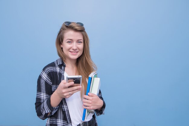 Studentin lokalisiert auf einem blauen Hintergrund mit Büchern, Notizbüchern und einem Smartphone in ihren Händen, die die Kamera betrachten