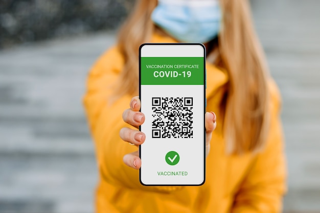 Studentin mit medizinischer Maske, die einen Handy-Bildschirm mit einer Anwendung zur Impfung gegen Coronavirus, Covid-19, Stadthintergrund, Kopierraum zeigt