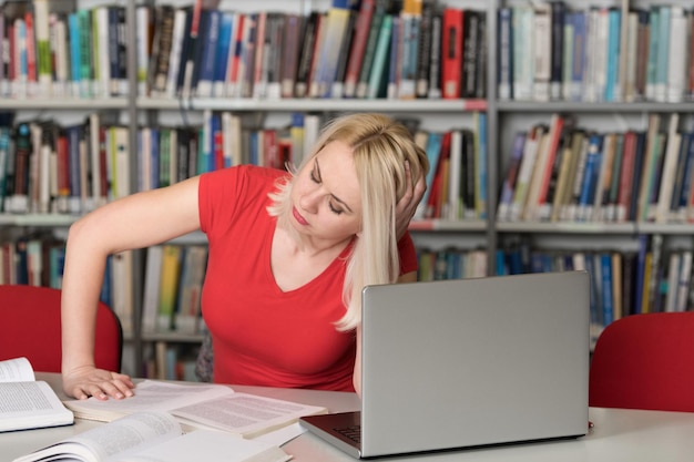 Studentin sieht müde aus, während sie mit einem Laptop und Lehrbüchern in der Bibliothek studiert