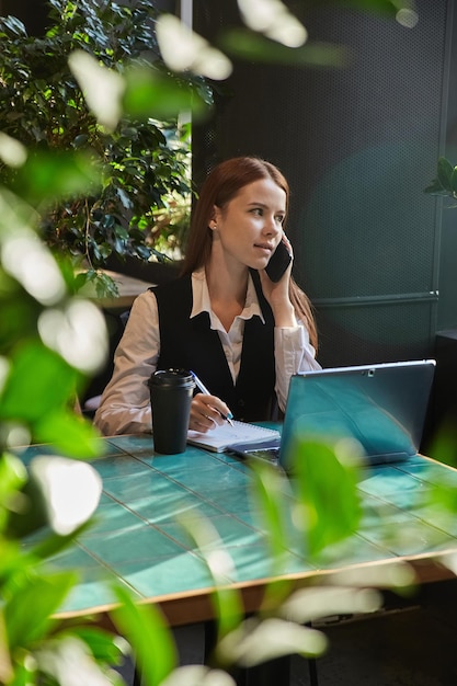 Studentin sitzt am Café-Tisch und lernt mit Laptop, der mit dem Smartphone anruft