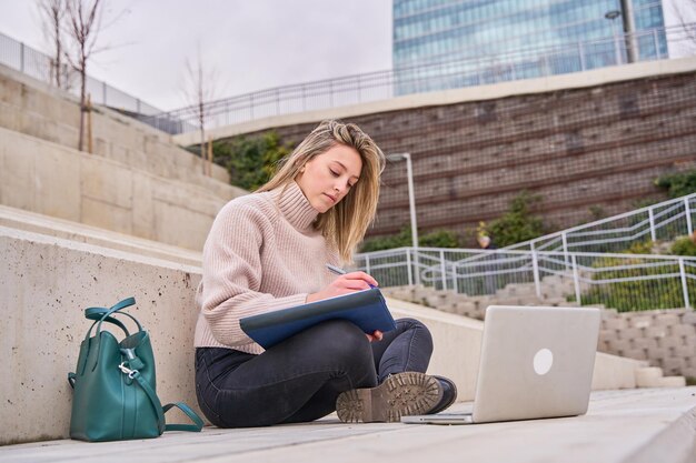 Studentin sitzt auf der Treppe und arbeitet am Laptop, bereitet sich auf Prüfungen im Freien vor und ruht sich auf dem Universitätscampus aus. Technologieausbildung und Fernarbeitskonzept