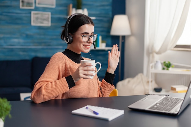 Studentin trägt Kopfhörer, während sie während eines Online-Videoanrufs mit ihrem College-Verräter winkt. Frau, die eine Tasse Kaffee hält und auf den Laptop schaut, der am Schreibtisch im Wohnzimmer sitzt