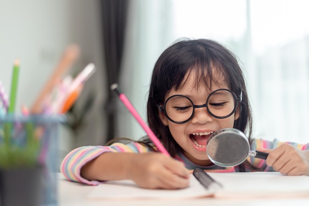 Studentisches kleines asiatisches Mädchen, das mit einer Lupe schreibt, die auf die Kamera schaut, süße Kinder, die im Wohnzimmer zu Hause sitzen, Familienaktivitätskonzept