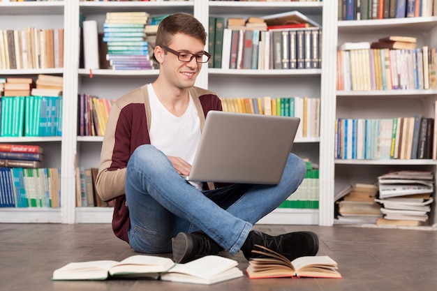 Studieren an einem ruhigen Ort. Fröhlicher junger Mann, der etwas auf dem Laptop tippt, während er auf dem Boden in der Bibliothek sitzt