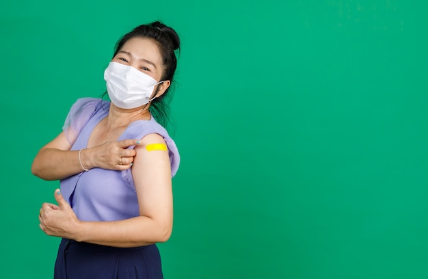 Studio Nahaufnahme einer asiatischen Patientin mit Gesichtsmaske Blick auf die Kamera, die den Daumen mit gelbem Verbandpflaster zeigt, nachdem sie vom Arzt auf grünem Hintergrund eine Coronavirus-Kovid-19-Impfung erhalten hat.