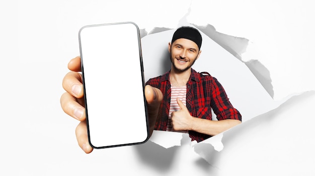 Studio-Porträt eines jungen lächelnden Mannes, der ein großes Smartphone mit Leerzeichen auf dem Bildschirm durch ein zerrissenes Papierloch zeigt Weißer Hintergrund Daumen nach oben mit rotem Hemd und schwarzem Band auf dem Kopf