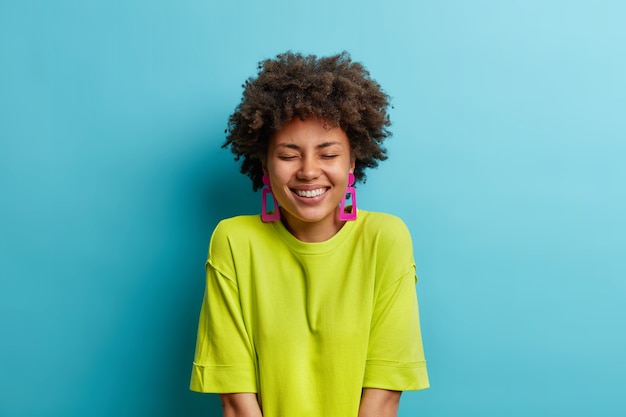 Studioaufnahme der positiven Frau mit Afro-Haar hält Augen geschlossen Lächeln vor Vergnügen zeigt weiße perfekte Zähne trägt lässiges grünes T-Shirt und Ohrringe isoliert über blaue Wand