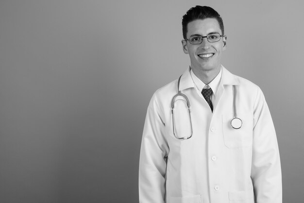 Studioaufnahme des jungen gutaussehenden Mannarztes, der Brillen gegen grauen Hintergrund in Schwarzweiss trägt