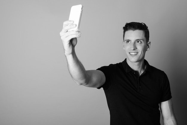 Studioaufnahme des jungen gutaussehenden Mannes, der Handy gegen grauen Hintergrund in Schwarzweiss verwendet