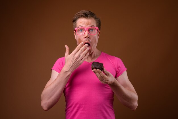 Studioaufnahme des jungen gutaussehenden Mannes, der rosa Hemd mit passenden rosa Brillen trägt, während Schokoladenkuchen gegen braunen Hintergrund haben