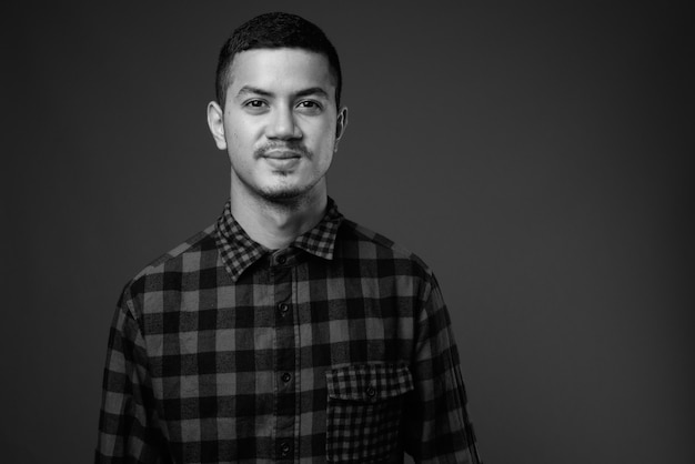 Studioaufnahme des jungen multiethnischen asiatischen Mannes, der kariertes Hemd gegen graue Wand in Schwarzweiss trägt