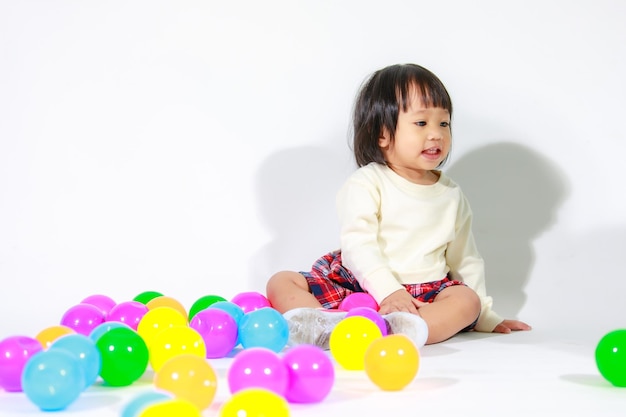 Studioaufnahme des kleinen niedlichen asiatischen Babymädchen-Tochtermodells mit kurzen schwarzen Haaren in lässigem kariertem Rock, das auf dem Boden sitzt und lachend mit bunten runden Kugeln spielt, spielt allein auf weißem Hintergrund.