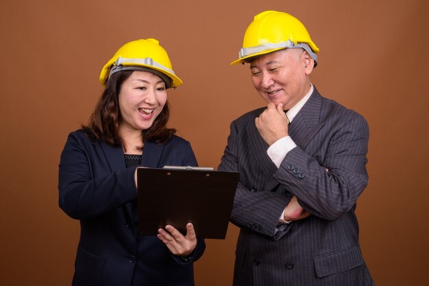 Studioaufnahme des reifen japanischen Geschäftsmannes und der reifen japanischen Geschäftsfrau, die Helm zusammen vor braunem Hintergrund tragen