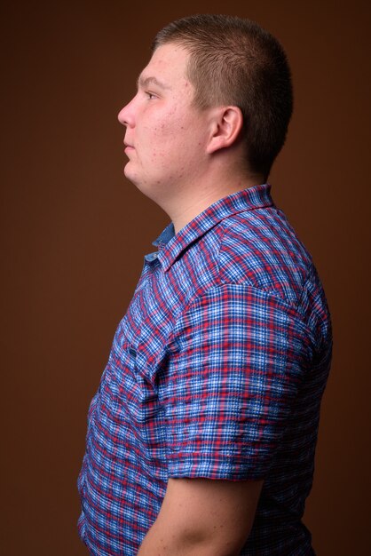Studioaufnahme des übergewichtigen jungen Mannes, der lila kariertes Hemd gegen braunen Hintergrund trägt