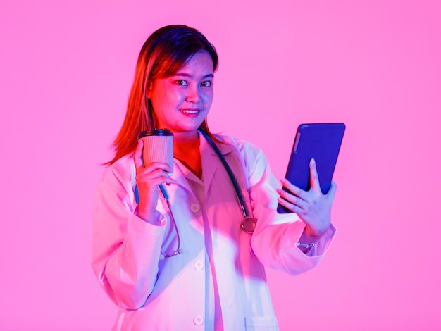 Studioaufnahme einer asiatischen erfolgreichen professionellen selbstbewussten Ärztin in Laborkitteluniform, die ein Stethoskop um den Hals hängt, eine Kaffeepause einlegen, die lächelnd einen Tablet-Computer auf rosafarbenem Hintergrund hält.
