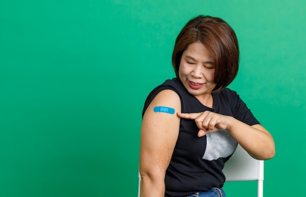 Studioaufnahme einer asiatischen Patientin mittleren Alters, die lächelt und einen blauen Gipsverband am Arm zeigt, nachdem sie von einem Arzt in der Klinik auf grünem Hintergrund eine Coronavirus-Kovid-19-Impfung erhalten hat.
