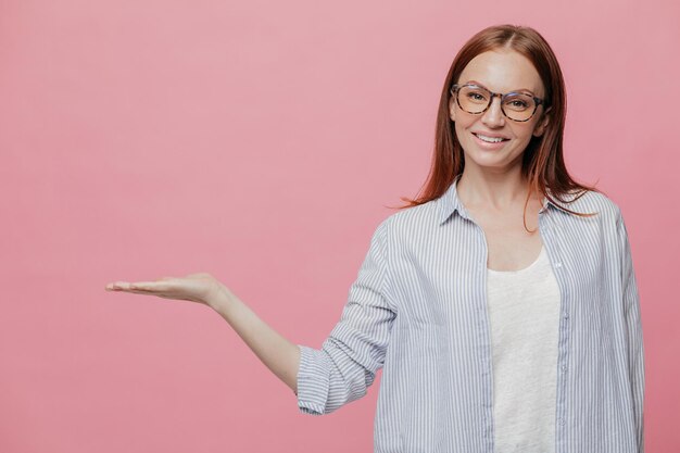 Studioaufnahme einer erfreuten kaukasischen Frau hält die Handfläche erhoben, trägt eine optische Brille, demonstriert etwas über dem Kopierraum, hält Leerzeichen, hat ein angenehmes Lächeln im Gesicht, posiert im Innenbereich über einer rosa Wand