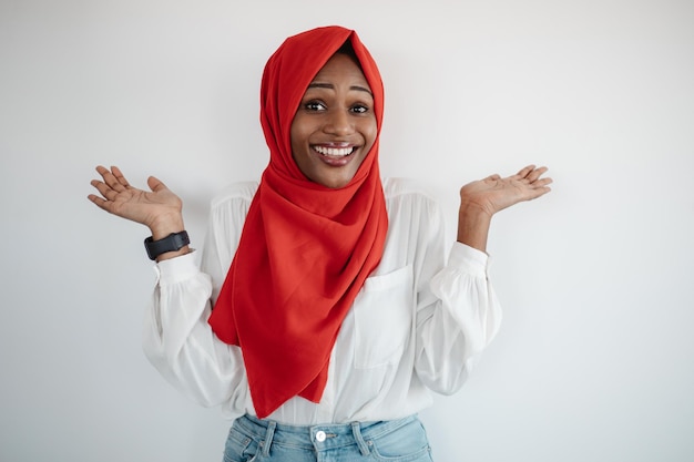 Studioaufnahme einer glücklichen schwarzen muslimischen Frau, die Handflächen ausbreitet, die Hände hochhebt und an der Kameralicht-Studiowand lächelt