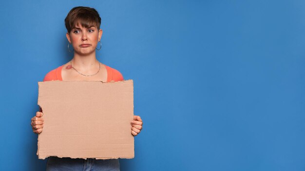 Studioaufnahme einer jungen Frau mit einem skeptischen Ausdruck, die ein leeres Pappblatt auf blauem Hintergrund hält. Kopieren Sie Platz, Platz für Ihre Anzeige oder Ihren Text.