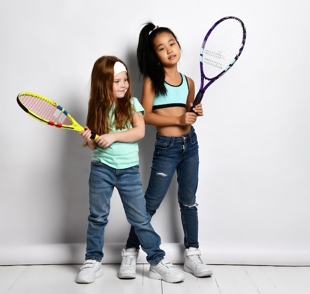 Studioaufnahme einer schönen Tennisspielerin im Teenageralter Kleine Freunde sind bereit, kaukasische und asiatische Kinder in modischer Kleidung mit einer Schlägerpose für die Kamera zu trainieren