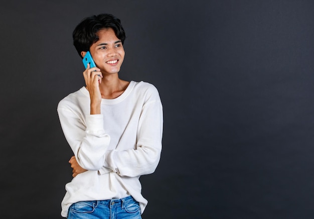 Studioaufnahme eines asiatischen jungen LGBTQ-Schwulen-Glamours, hübsches bisexuelles homosexuelles männliches Model in lässigem Outfit, das lächelnd mit Smartphone auf schwarzem Hintergrund steht