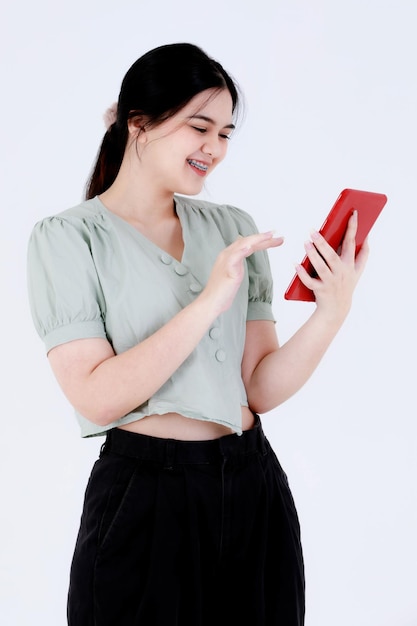 Studioaufnahme eines asiatischen jungen, schönen Teenager-Frauenmodells mit Pferdeschwanzfrisur, das ein lässiges Crop-Top-Outfit trägt, das mit berührendem Touchscreen-Tablet in rotem Gehäuse auf weißem Hintergrund steht.