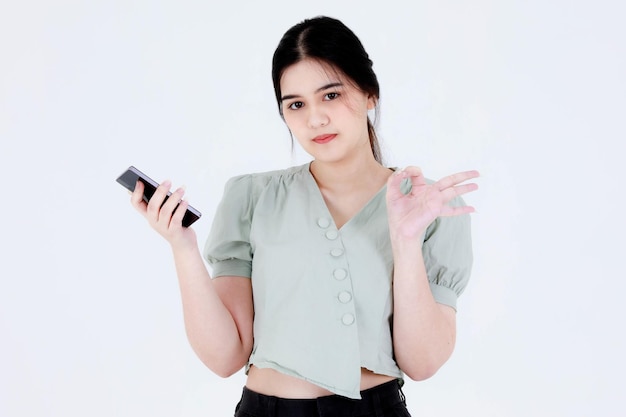 Studioaufnahme eines asiatischen jungen Teenager-Frauenmodells mit Pferdeschwanzhaaren, das Zahnpflege-Zahnspangen und ein bauchfreies Outfit trägt, das mit winkender Hand grüßt, sagt Hallo auf Smartphone-Videoanruf auf weißem Hintergrund.