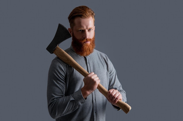 Studioaufnahme eines brutalen Kerls mit rotem Bart Brutaler Kerl mit Bartgriffaxt isoliert auf grauem Hintergrund