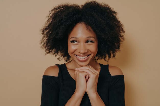 Studioaufnahme eines glücklichen Afro-Mädchens hält die Hände unter dem Kinn zusammen, ist in Hochstimmung, mag die Idee von Freunden für den nächsten Tag, einzeln auf dunkelbeigem Wandhintergrund mit Kopierraum. Konzept der positiven Emotionen