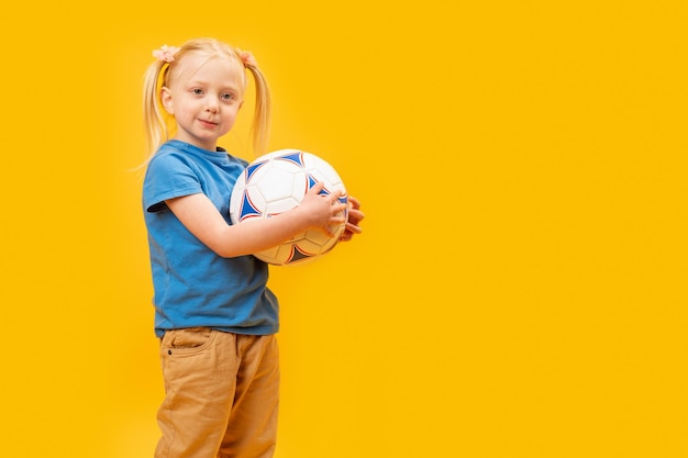 Studioaufnahme eines jungen blonden Mädchens, das einen Fußball vor gelbem Hintergrund hält Kind und Fußball-Kopierraum-Attrappe