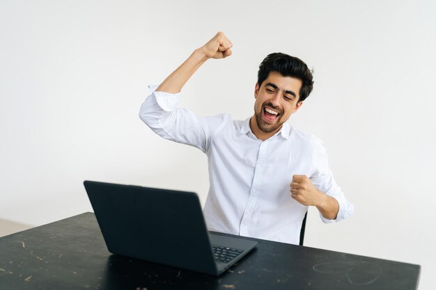 Studioaufnahme eines lächelnden, aufgeregten männlichen Büroangestellten, der Eureka schreit und die Hände hochhebt, um eine schwierige Aufgabe zu lösen, die am Schreibtisch auf weißem, isoliertem Hintergrund sitzt