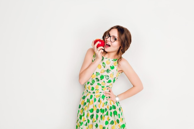 Studioaufnahme eines lächelnden Brunettemädchens, das weit lächelt. Mädchen trägt eine schwarze Brille und ein gelbes und grünes Kleid. In der rechten Hand hält sie einen roten Apfel und will darauf beißen.