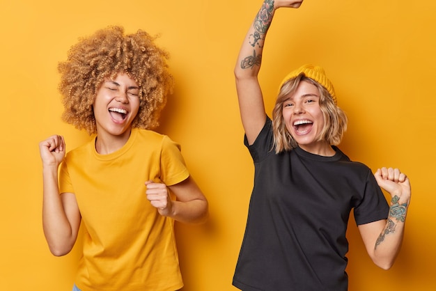 Studioaufnahme von fröhlichen, lebhaften jungen Frauen, die das Leben genießen, die Arme schütteln, sich energisch ausrufen, laut lässige Basic-T-Shirts tragen, die über gelbem Hintergrund isoliert sind, Spaß haben und tanzen Fröhliches Gefühlskonzept