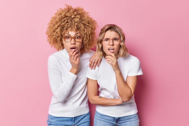 Studioaufnahme von schockierten jungen Frauen, die staunend starren, können nicht glauben, dass ihre Augen vor Überraschung den Atem anhalten, gekleidet in lässige weiße T-Shirts, die über rosafarbenem Hintergrund isoliert sind Konzept der menschlichen Reaktionen