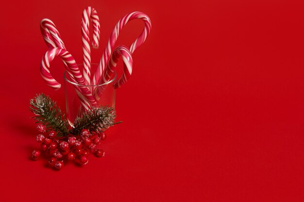 Studioaufnahme von schöner minimalistischer einfacher Komposition mit Weihnachtslutschern in transparentem Glas und schneebedecktem Kiefernzweig mit roten Beeren, Stechpalme, auf rotem Hintergrund mit Kopienraum für Werbung