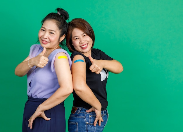 Studioaufnahme von zwei asiatischen, glücklichen Patientinnen mittleren Alters, die lächelnd auf die Kamera blicken, die den Daumen mit Verbandpflaster zeigt, nachdem sie den Coronavirus-Kovid-19-Impfstoff auf grünem Hintergrund erhalten haben.