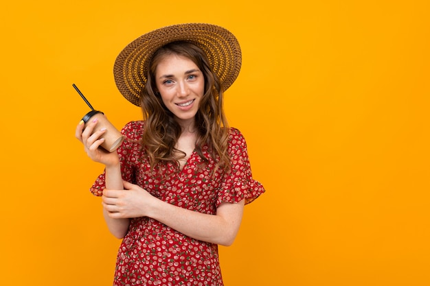 Studiocharming elegante niña sonriente con un sombrero y un vestido rojo con un vaso de papel sobre una pared naranja