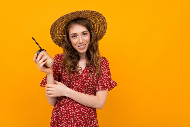 Studiocharming menina sorridente elegante com um chapéu e um vestido vermelho com um copo de papel em uma parede laranja