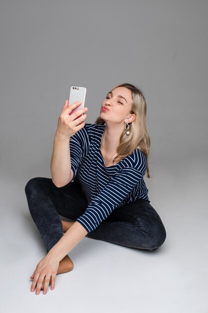 Studiofoto eines hübschen jungen blonden Mädchens in Freizeitkleidung, das auf dem Boden sitzt und Selfie mit schmollenden Lippen macht.