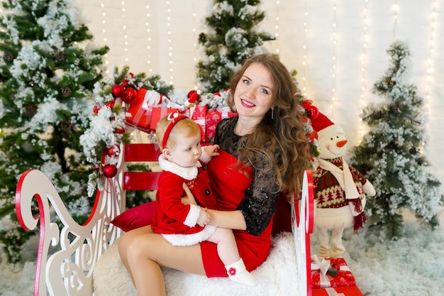 Studioporträt der glücklichen Mutter und der Tochter in den Kleidern des Weihnachtsmanns, die nahe Weihnachtsbaum sitzen