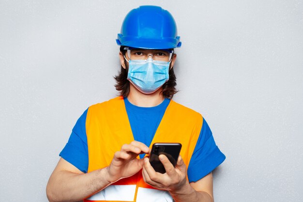 Studioporträt des jungen Arbeiteringenieurs unter Verwendung des Smartphones, das medizinische Gesichtsmaske gegen Coronavirus oder covid-19 trägt